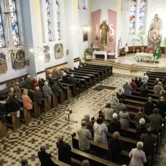 Ludzie siedzą w ławkach podczas mszy wieczornej przed świętem. Witraże i obrazy zdobią wnętrze kościoła.